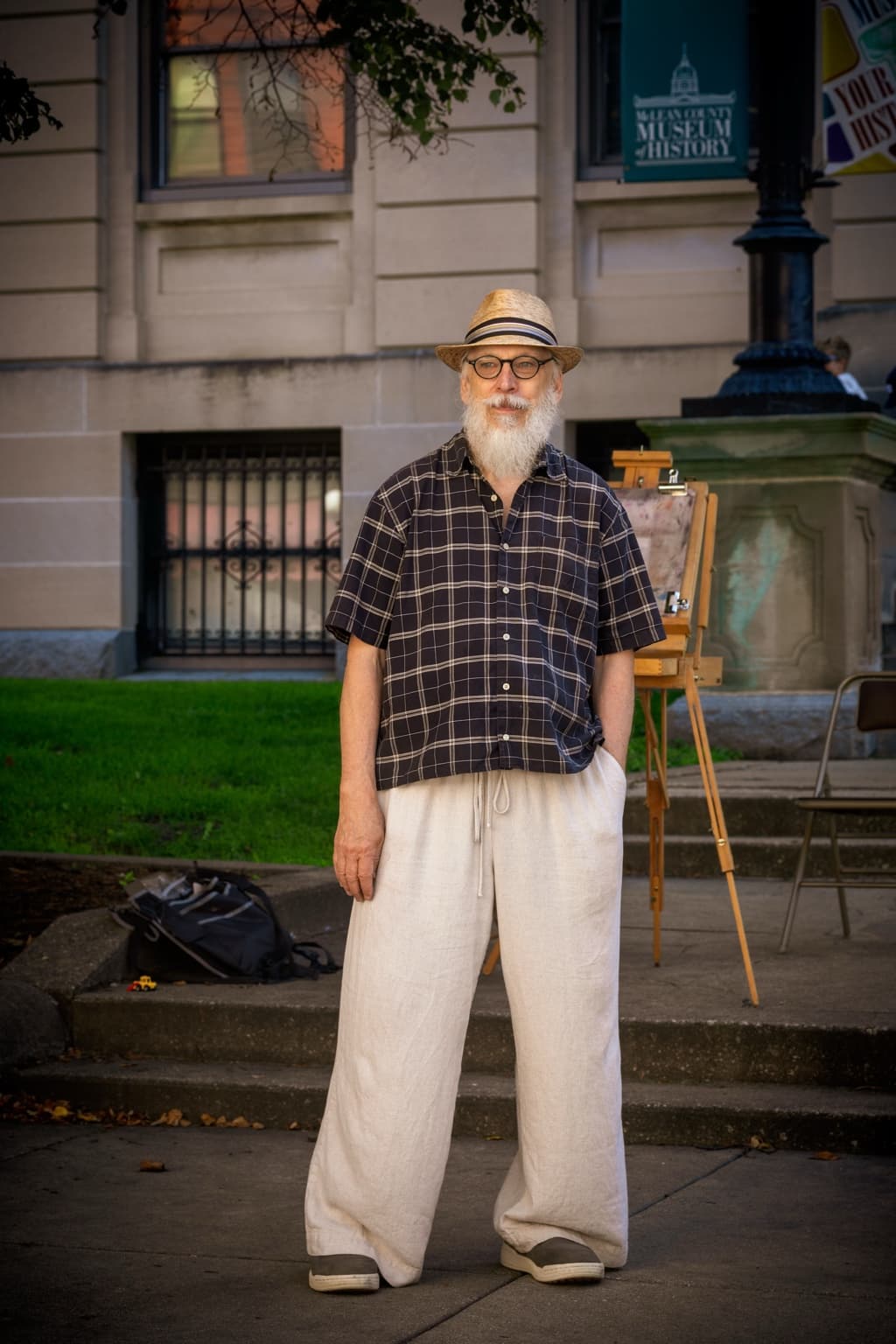 Artist Brian K. Simpson stands beside his easel outside the McLean County Museum of History wearing a straw hat and round glasses.