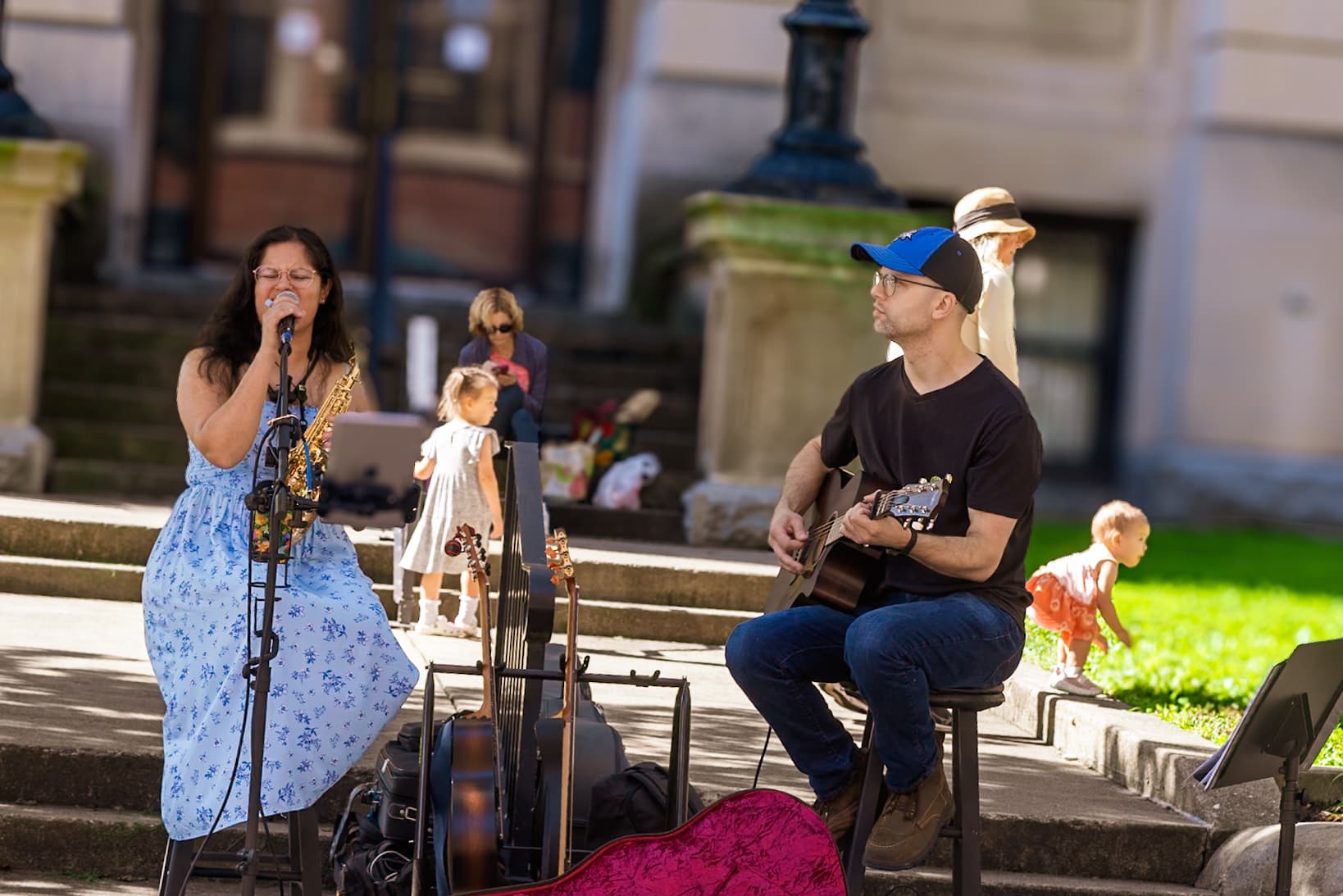 Micro Spice performs on the museum steps with vocals, saxophone and acoustic guitar as families move through the background.