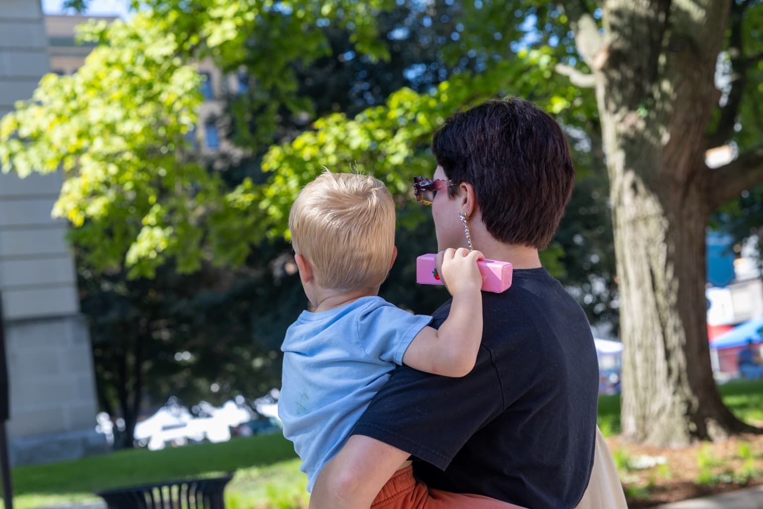An adult carries a young child beneath the trees near the Downtown Bloomington Farmers Market.